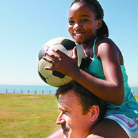 girl holding soccer ball sitting on top of man's shoulders