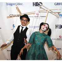 Man in suit and bowler hat and woman in green dress stand with strings attached to their arms, in costume as marionette puppets