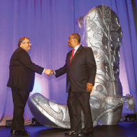 Jorge de la Riva and Val Bias shake hands onstage with large silver cowboy boot behind