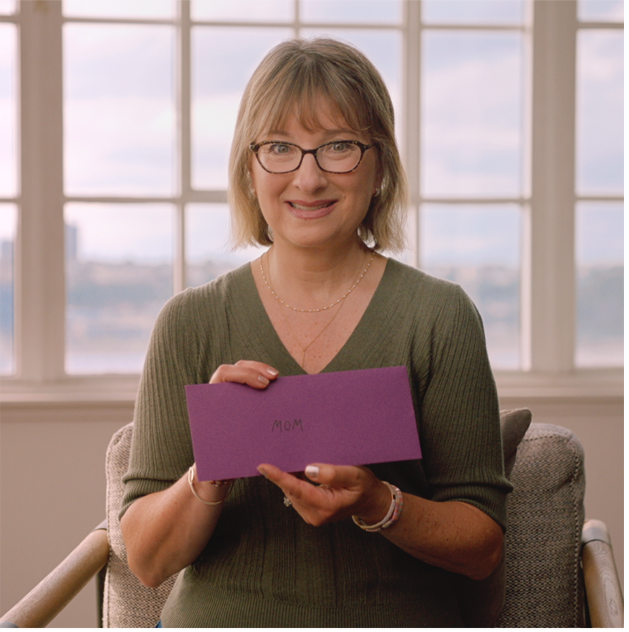 Suzanne smiling holding an envelope with a card inside that says Mom.