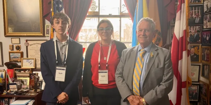 Two advocates, pose with an older man in a suit and tie inside a legislative office decorated with flags and framed photos, during a Washington Days advocacy visit.