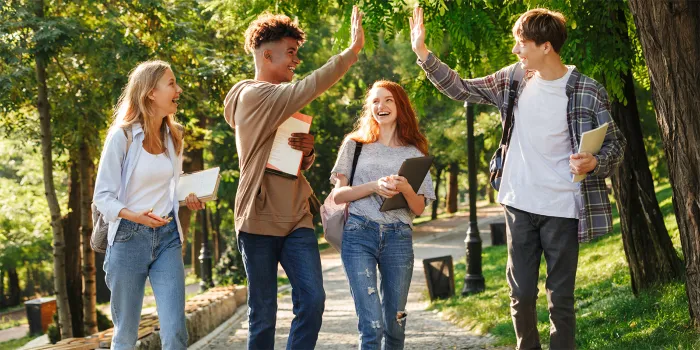 A group of four teenagers smiling and walking in a park together holding notebooks. Two of the boys are giving each other a high-five