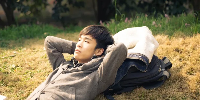 Man lies back in the grass with his hands behind his head, resting with his jacket and backpack beside him.
