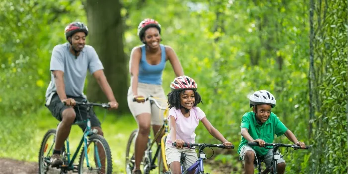 A family of four wearing helmets rides bicycles together on a wooded trail, smiling and enjoying an active outing.