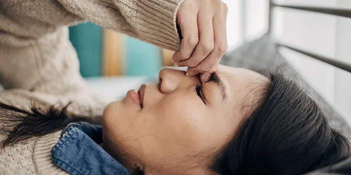 A woman lying down on her bed with a headache while holding the bridge of her nose