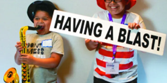 Two kids - one playing toy saxophone, other in striped red hat with sign "Having a Blast"