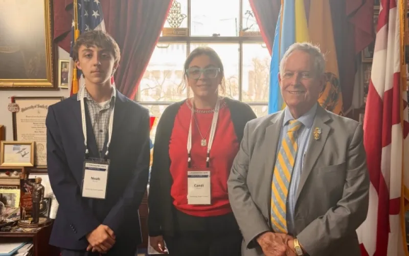 Two advocates, pose with an older man in a suit and tie inside a legislative office decorated with flags and framed photos, during a Washington Days advocacy visit.