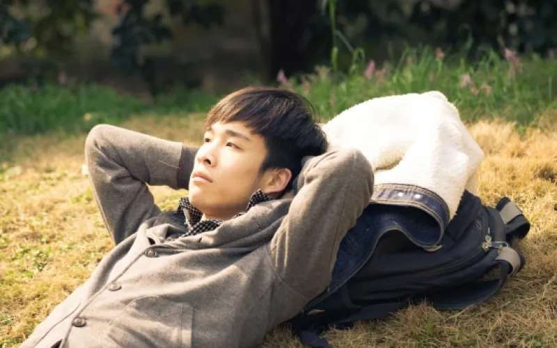 Man lies back in the grass with his hands behind his head, resting with his jacket and backpack beside him.