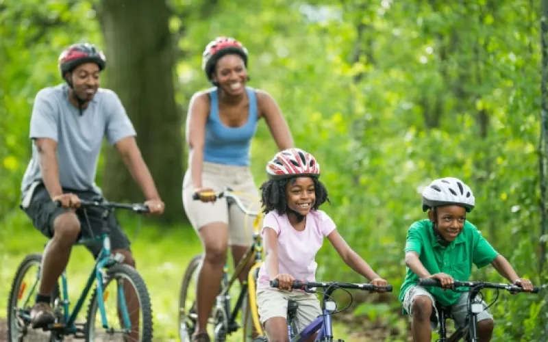 A family of four wearing helmets rides bicycles together on a wooded trail, smiling and enjoying an active outing.