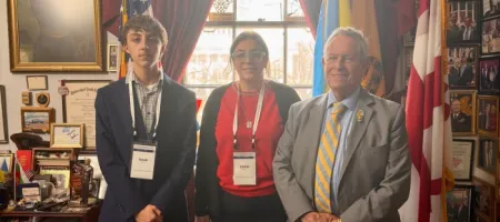 Two advocates, pose with an older man in a suit and tie inside a legislative office decorated with flags and framed photos, during a Washington Days advocacy visit.