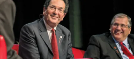 Close up of man in gray suit and red tie, sitting onstage with other panelists