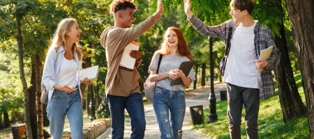 A group of four teenagers smiling and walking in a park together holding notebooks. Two of the boys are giving each other a high-five