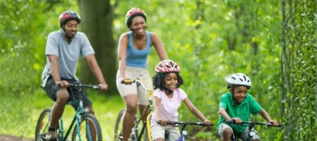 A family of four wearing helmets rides bicycles together on a wooded trail, smiling and enjoying an active outing.