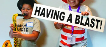 Two kids - one playing toy saxophone, other in striped red hat with sign "Having a Blast"