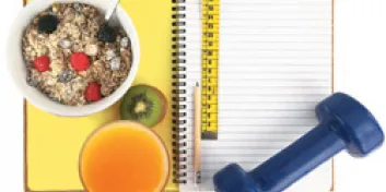 Bowl of granola with fruit and juice on top of open lined notebook with yellow measuring tape, pencil and blue hand weight